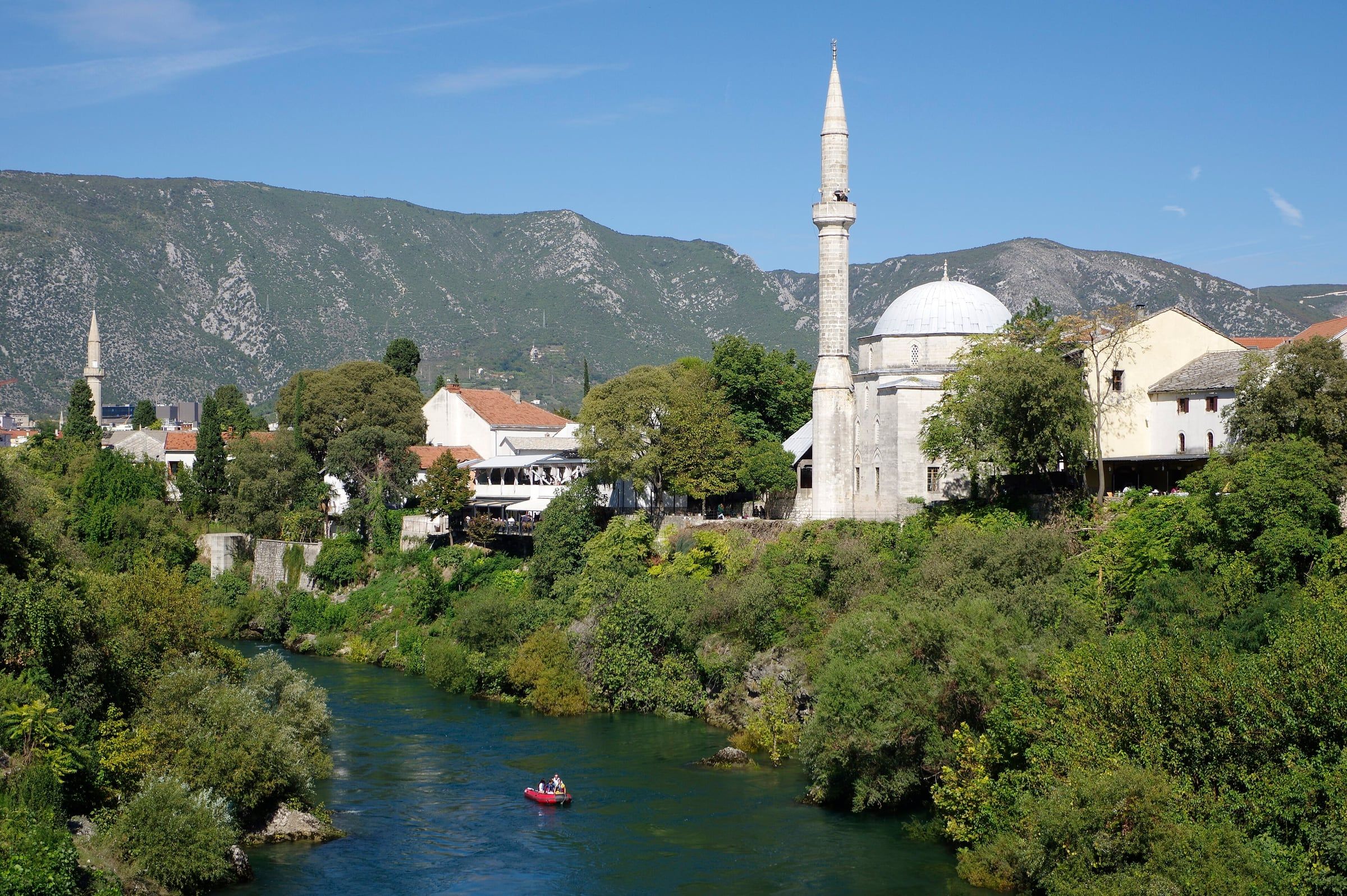 The white dome and slender minaret of the Koski Mehmed Pasha Mosque above trees on the bank of the Neretva, with kayaks on the river.