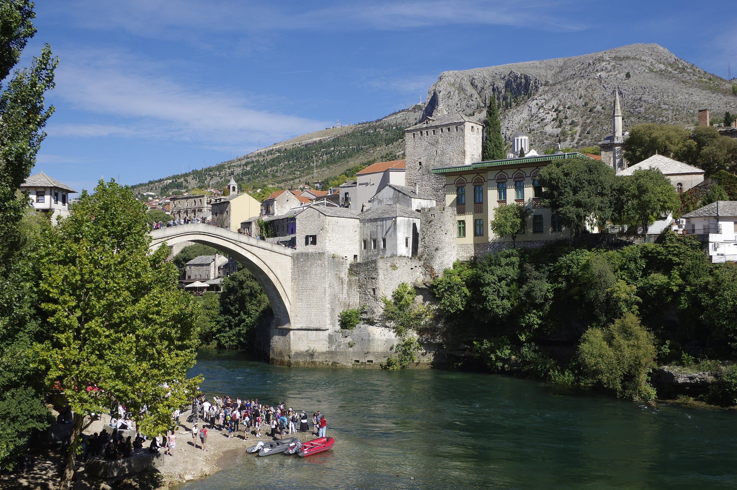 The single stone arch of the Old Bridge spanning the green Neretva, with stone houses and a minaret on the bank and a bare mountain behind.